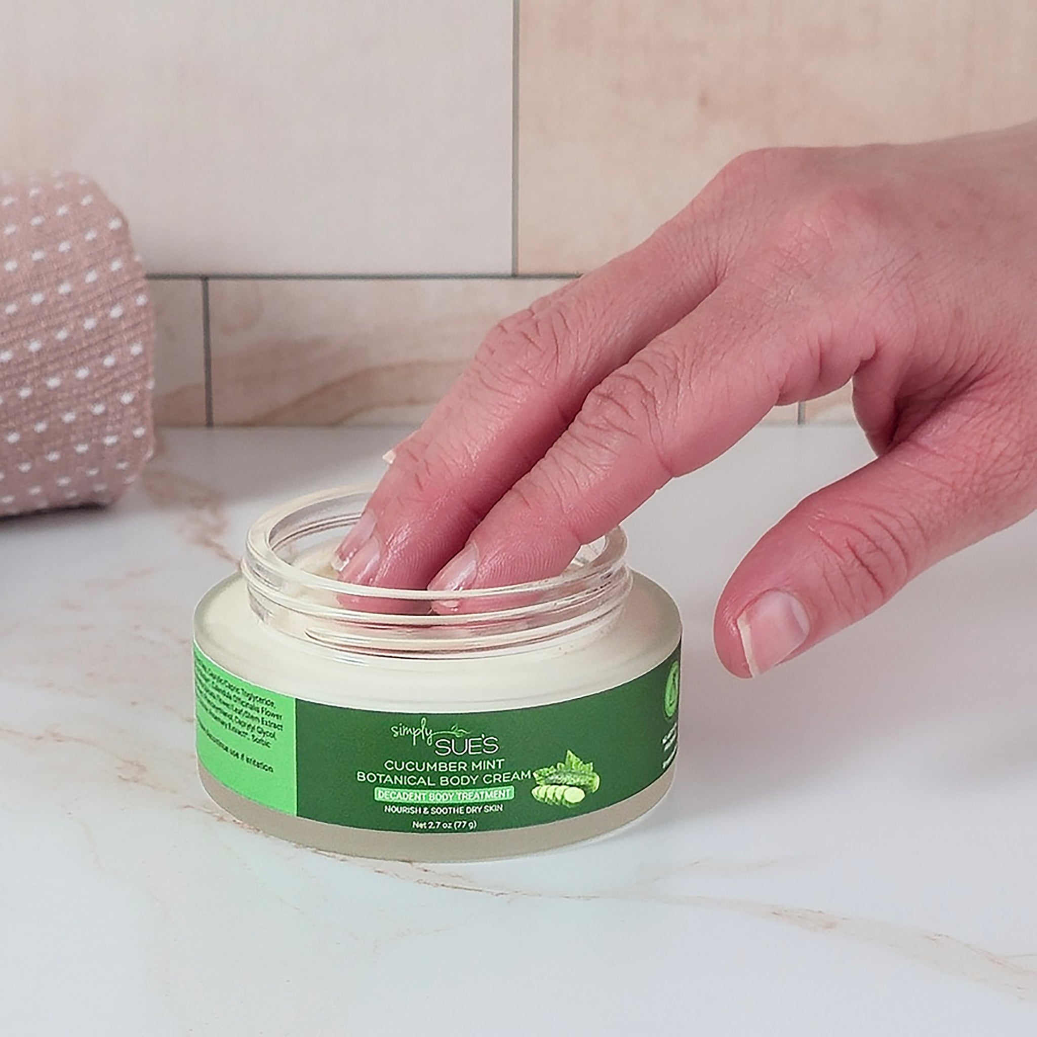 Hand opening a jar of Simply Sues cucumber mint botanical soothing cream on a bathroom counter.