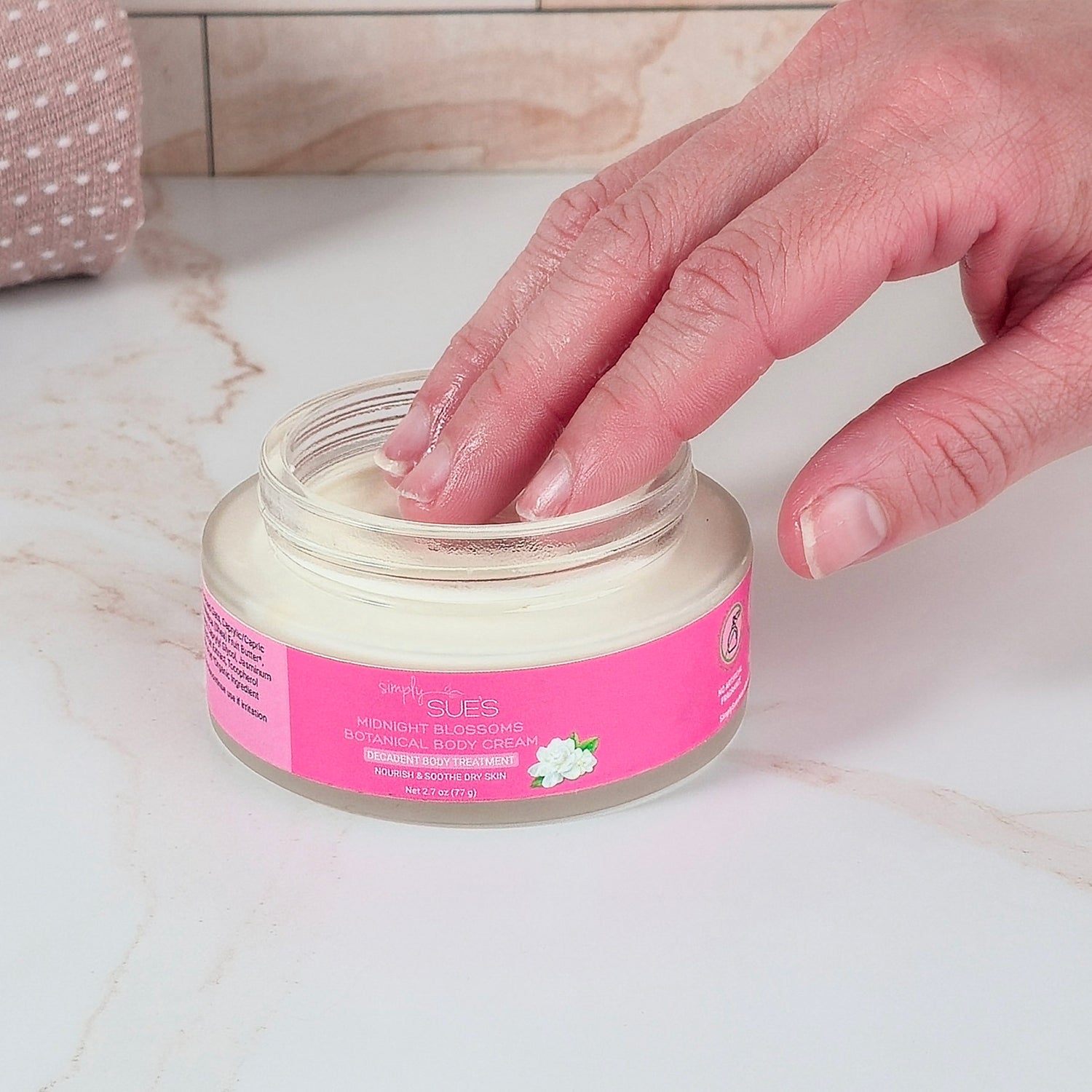 Hand applying cream from a jar with a pink label on a marble surface