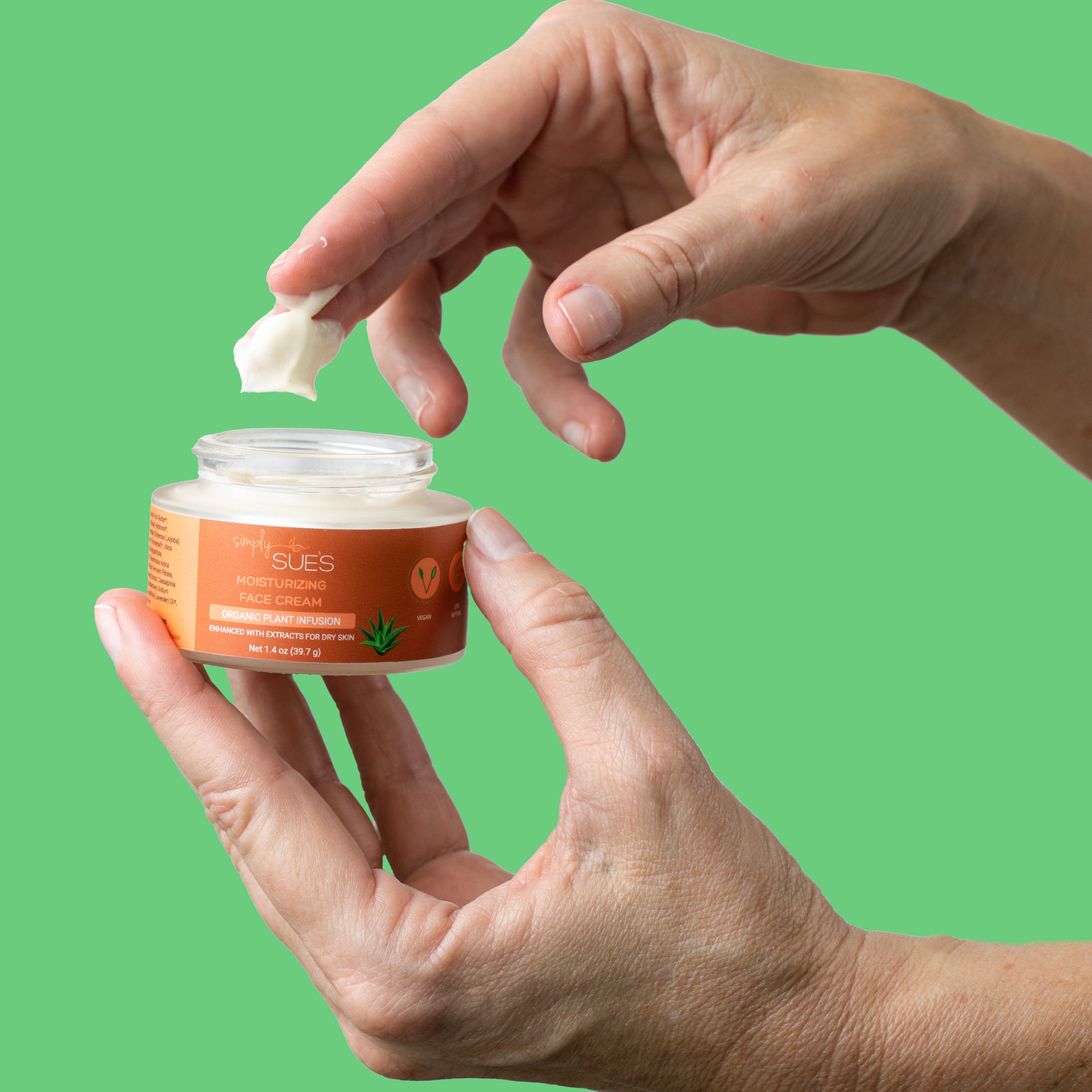 A hand holding an open jar of Simply Sue's Moisturizing Face Cream with cream on her fingers to show the texture